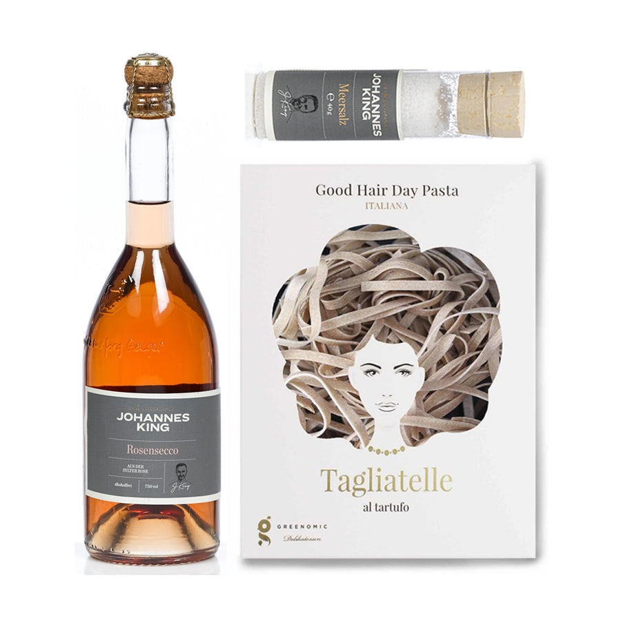 Das „Geschenk mit Liebe und Tartufo“ der Sylter Manufaktur Johannes King ist elegant auf weißem Hintergrund arrangiert. Dieses Gourmet-Geschenk besteht aus einer Flasche Johannes King Rosésecco, einem Zylinder Sommertrüffel Rosé-Salz und einer Schachtel Good Hair Day Pasta Tagliatelle al Tartufo, die mit einer Illustration eines Frauenkopfes aus Tagliatelle verziert ist.