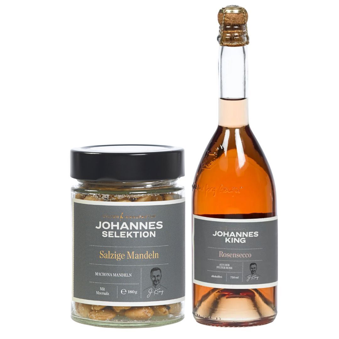 Ein Glas mit gesalzenen Marcona-Mandeln mit der Aufschrift „Salted Almonds“ wird mit einer Flasche Rosé-Sekt Sylter Heckenrose serviert. Beide Produkte stammen aus der Linie Sylter Manufaktur Johannes King und verfügen über elegante schwarze Etiketten mit Porträtskizzen, die einen Hauch von Raffinesse und Stil ausstrahlen.