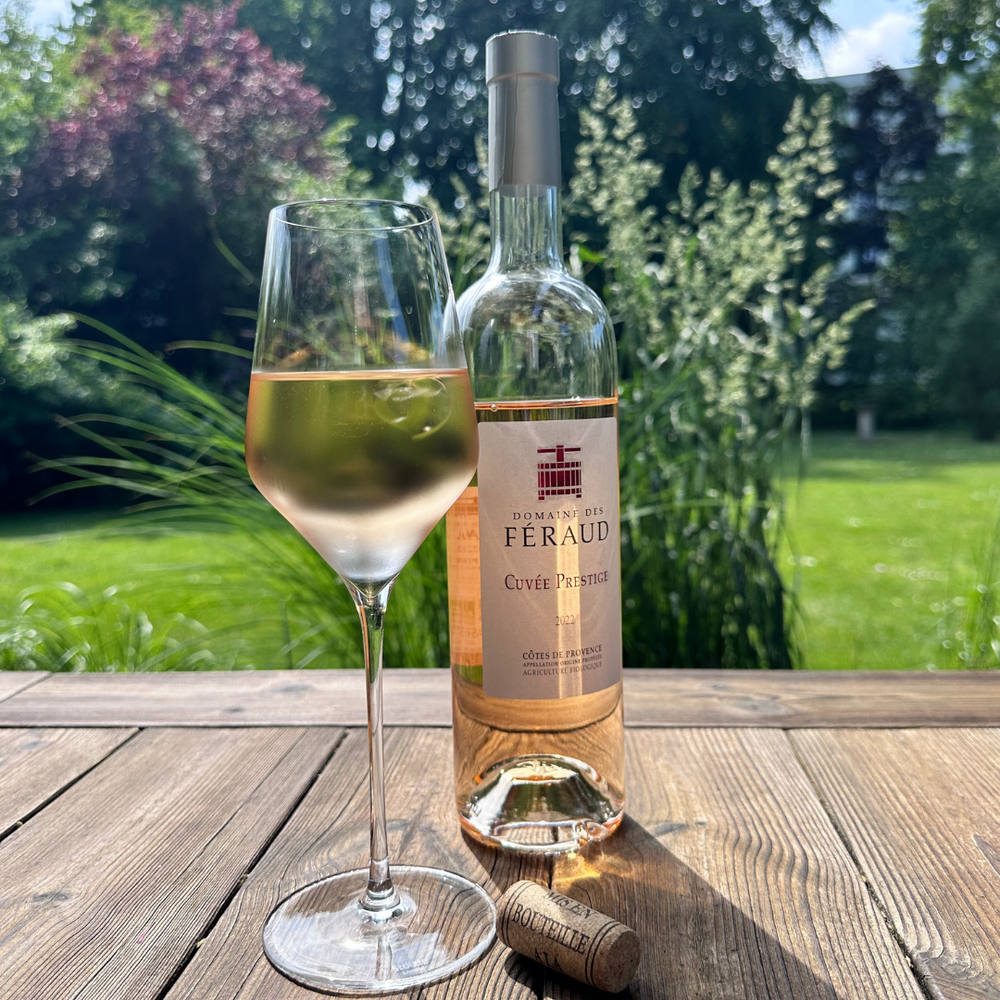 Eine Flasche Roséwein 2022 Domaine des Féraud - Cuvée Prestige steht neben einem gefüllten Weinglas auf einem Holztisch. Ein Korken mit dem Text „Domaine des Féraud“ liegt daneben. Der Hintergrund ist ein grüner Garten, in dem Sonnenlicht durch Bäume und Sträucher fällt.