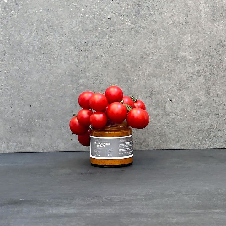 Ein Behälter mit Mojo Rojo-Sauce der Sylter Manufaktur steht auf einer dunklen Fläche vor einem grauen Steinhintergrund. Ein Bündel roter Kirschtomaten ist auf dem Behälter angeordnet und hebt ihn als perfekten Dip für Mojo-Sauce hervor.