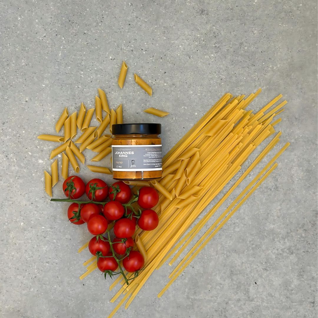 Auf einem grauen Untergrund sind rohe Penne und Spaghetti in Herzform ausgelegt. Im Inneren des Herzens befinden sich eine Traube Strauchtomaten und ein Glas „Pasta mit Mojo Rojo“ mit der Aufschrift „Sylter Manufaktur Johannes King“.