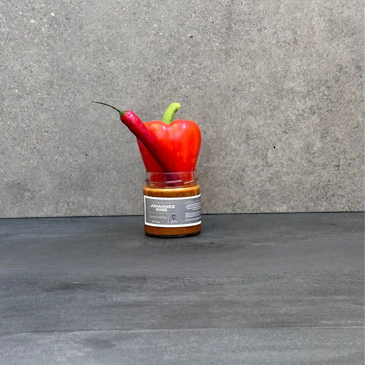 Ein kleines Gefäß mit der Aufschrift „SYLTER MANUFAKTUR“ steht auf einer dunklen Fläche vor grauem Hintergrund. Eine rote Paprika und eine rote Chilischote balancieren darauf und ergeben ein kunstvolles Arrangement. Dies könnte das inspirierende Herzstück Ihres nächsten Dip-Trios sein, mit Geschmacksrichtungen wie Paprika-Chili-Dip oder Mojo Rojo.