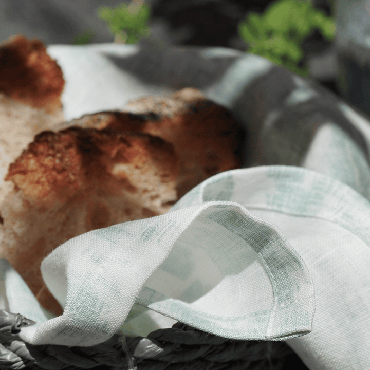 Eine Nahaufnahme eines Brotkorbs mit einem 4er-Set Servietten von Dorothea Waydsch - Minze, in dem Stücke von handwerklich hergestelltem Brot liegen. Das Brot hat eine knusprige, gebräunte Kruste und im Hintergrund ist verschwommenes Grün zu sehen.