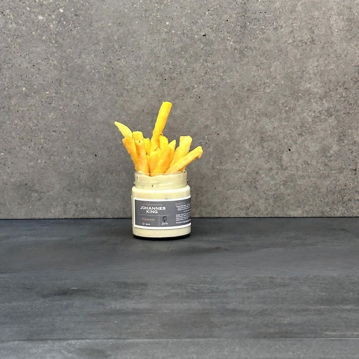 Ein kleines Glasgefäß mit Joannes Aioli-Sauce ist mit einem Haufen goldbrauner Pommes Frites bedeckt. Das Glas mit der Aufschrift „Sylter Dip-Duo“ von Sylter Manufaktur steht auf einer dunklen Oberfläche mit einer grauen, strukturierten Wand im Hintergrund.