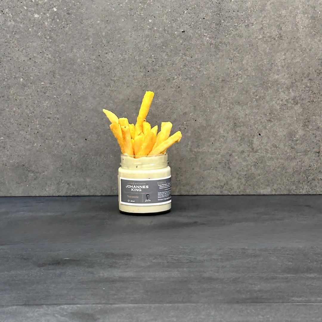 Ein kleines Glas mit der Aufschrift „Sylter Manufaktur Sylter Dip-Set“ ist mit Pommes Frites gefüllt und erinnert mit seiner Gourmet-Note an eine Kreation von Johannes King. Das Glas steht auf einer dunklen Fläche vor einem grauen, strukturierten Hintergrund.