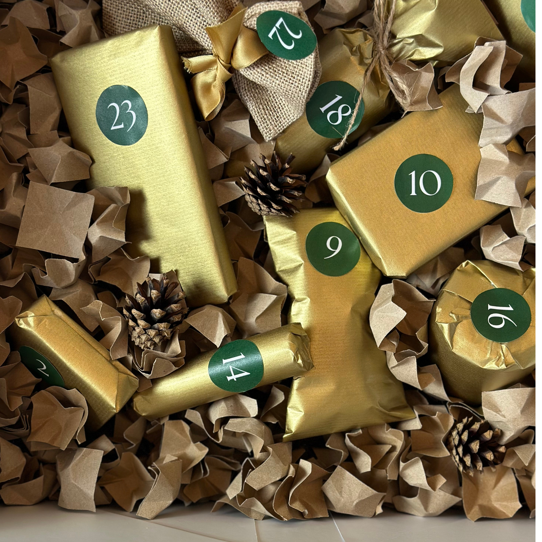 Ein Arrangement aus goldverpackten Geschenken aus dem Adventskalender 2024 der Sylter Manufaktur Johannes King, mit grünen nummerierten Aufklebern für die Tage 9, 10, 12, 14, 16, 18 und 23. Diese sind wunderschön auf zerknülltem braunem Papier präsentiert, zwischen denen Tannenzapfen eingestreut sind.