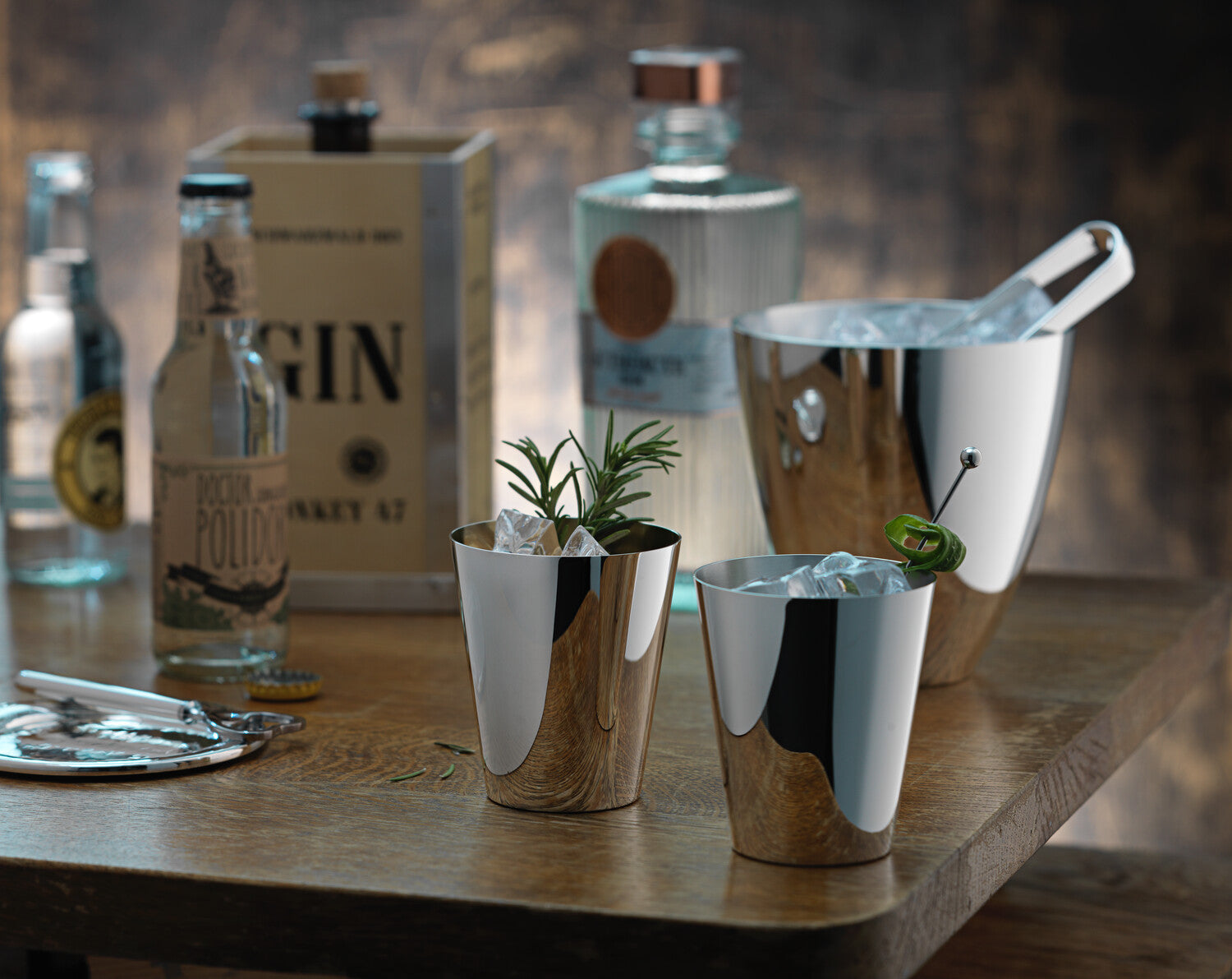 Eine Auswahl an Ginflaschen mit einem Eiskübel aus Edelstahl und zwei modernen, mit Eis gefüllten Cocktailgläsern auf einem Holztisch.