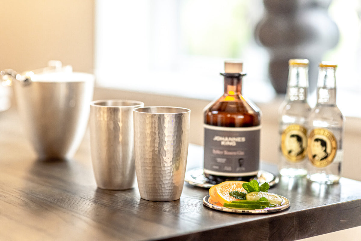 Ein ordentlich gedeckter Tisch mit Cocktailzutaten, darunter zwei Metallbecher, eine Flasche handwerklich hergestellter Sirup und zwei Flaschen Tonic Water, dazu ein Teller mit geschnittenen Zitrusfrüchten.