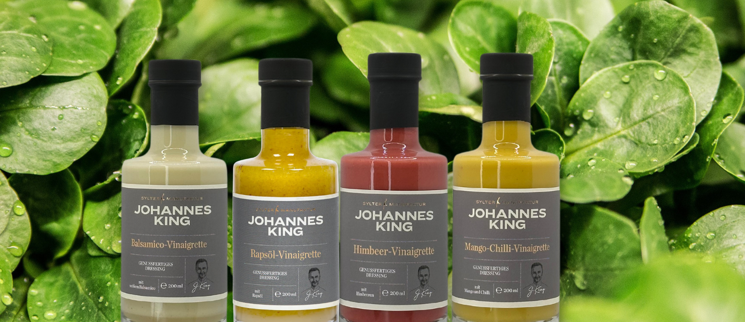 Ein Satz von vier Salatdressingflaschen der Marke Johannes King, die in einer Reihe vor einem Hintergrund aus frischen grünen Blättern mit Wassertropfen angeordnet sind.
