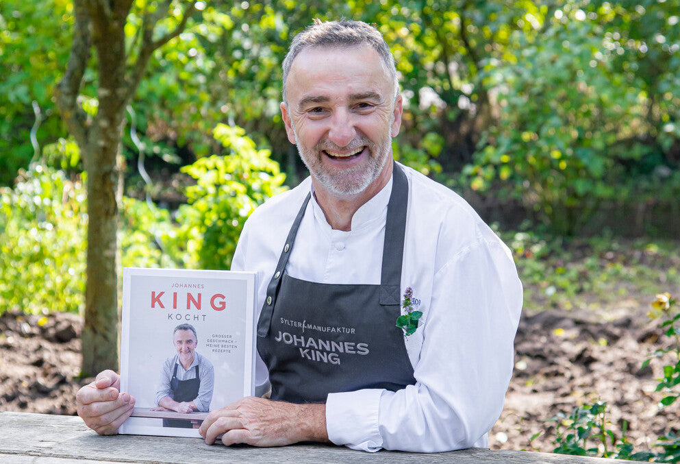 Ein lächelnder Mann mit Kochschürze zeigt ein Kochbuch, dessen Titel seinen Namen enthält.