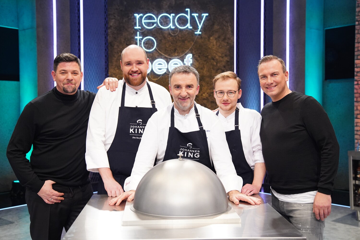 Fünf Männer stehen hinter einer Theke mit einer großen metallischen Kuppelabdeckung in der Küche eines Fernsehstudios und scheinen Teil eines Kochwettbewerbs oder einer Kochshow zu sein.
