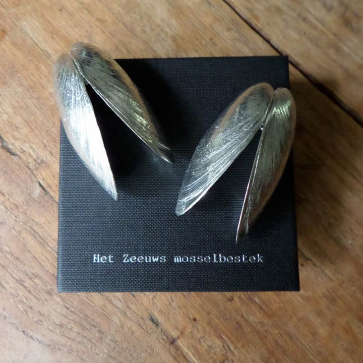 Eine schwarze quadratische Karte mit silbernen muschelförmigen Utensilien von der Sylter Manufaktur Johannes King, genannt Muscheln-Besteck. "Het Zeeuws mosselbestek" ist auf die Karte gedruckt, die auf einer hölzernen Unterlage ruht.