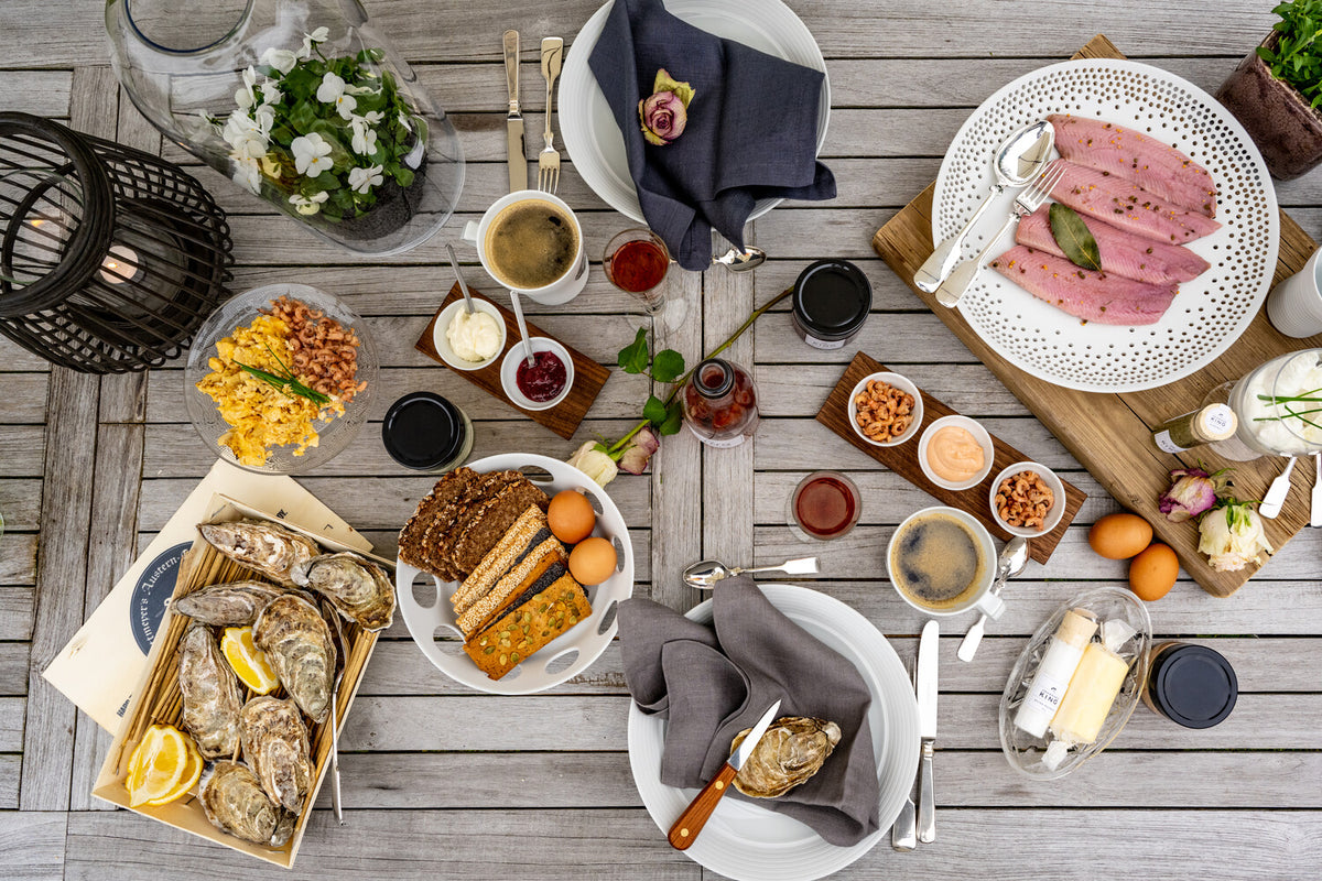 Ein übersichtlicher Esstisch im Freien mit einer Auswahl an Speisen, darunter Austern, Fleischscheiben, Gewürzen und Brot, mit ordentlich platziertem Besteck und Servietten.