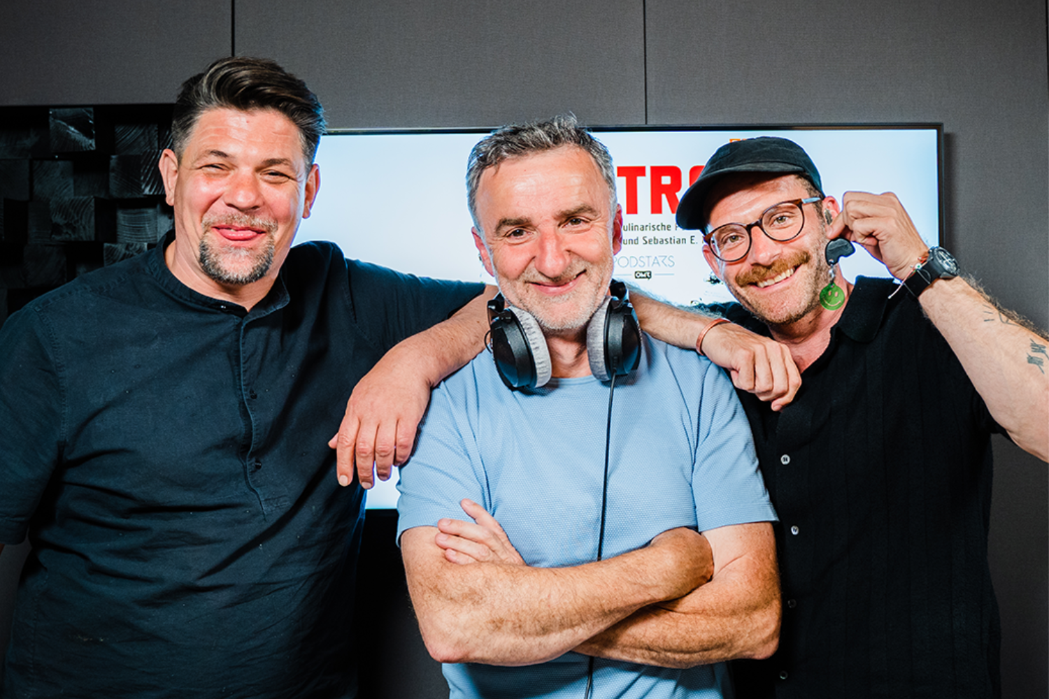 Drei lächelnde Männer in einem Studio, einer von ihnen trägt Kopfhörer um den Hals.