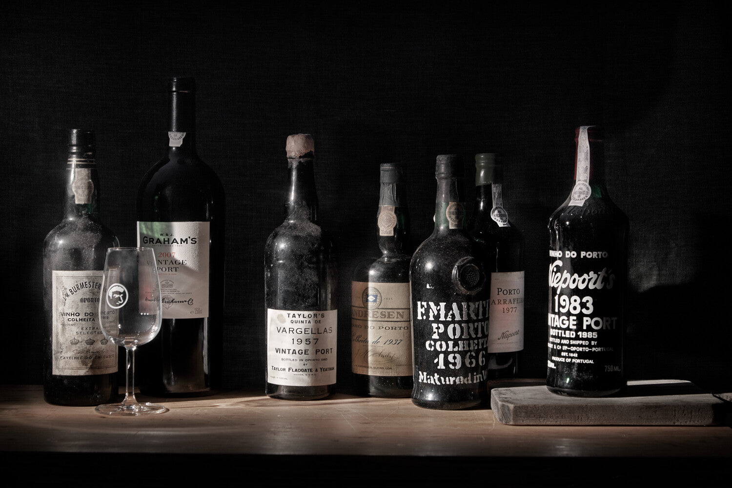 Eine Sammlung alter Portweinflaschen und ein Glas auf einem Tisch vor dunklem Hintergrund.