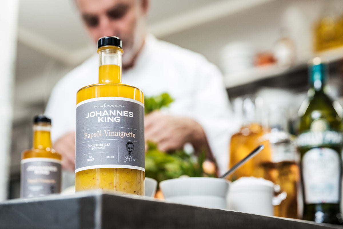 Im Vordergrund steht eine Flasche Rapsöl-Vinaigrette von Johannes King, während im Hintergrund ein verschwommener Koch in einer professionellen Küche Speisen zubereitet.