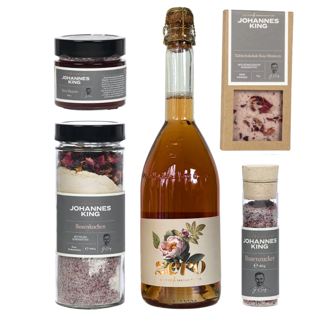 Das Set "Rosige Zeiten - Groß" der Sylter Manufaktur Johannes King enthält Rosenmarmelade, ein blumiges Sektgetränk, Schokolade, Backmischung und eine Rosenzuckermühle, alle mit grauen Johannes King-Etiketten.