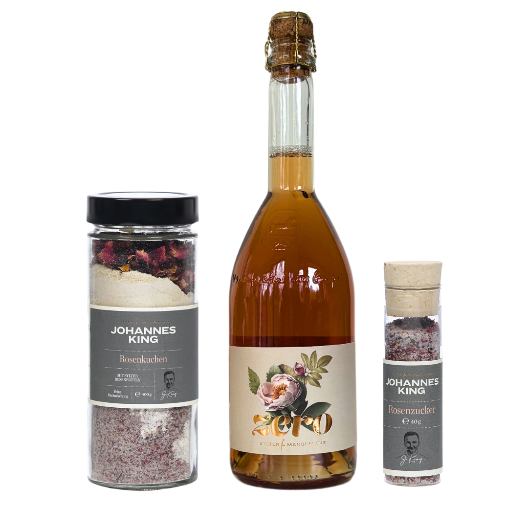 Eine klare Flasche prickelndes Getränk mit einem Blumenetikett steht zwischen zwei Gläsern mit der Aufschrift "Süße rosige Verführung von der Insel - Groß" der Sylter Manufaktur Johannes King, alles auf weißem Hintergrund.