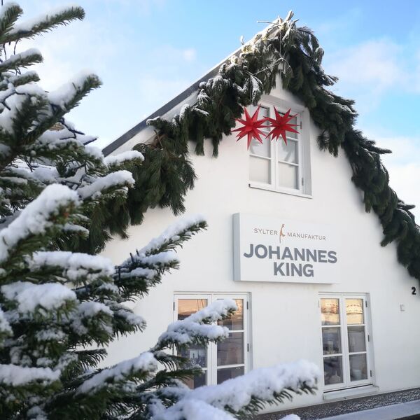 Ein schneebedecktes Gebäude mit einem Schild mit der Aufschrift „Johannes King“ ist mit einer großen roten Sterndekoration geschmückt und liegt eingebettet zwischen immergrünen Bäumen, die ebenfalls mit Schnee bestäubt sind.