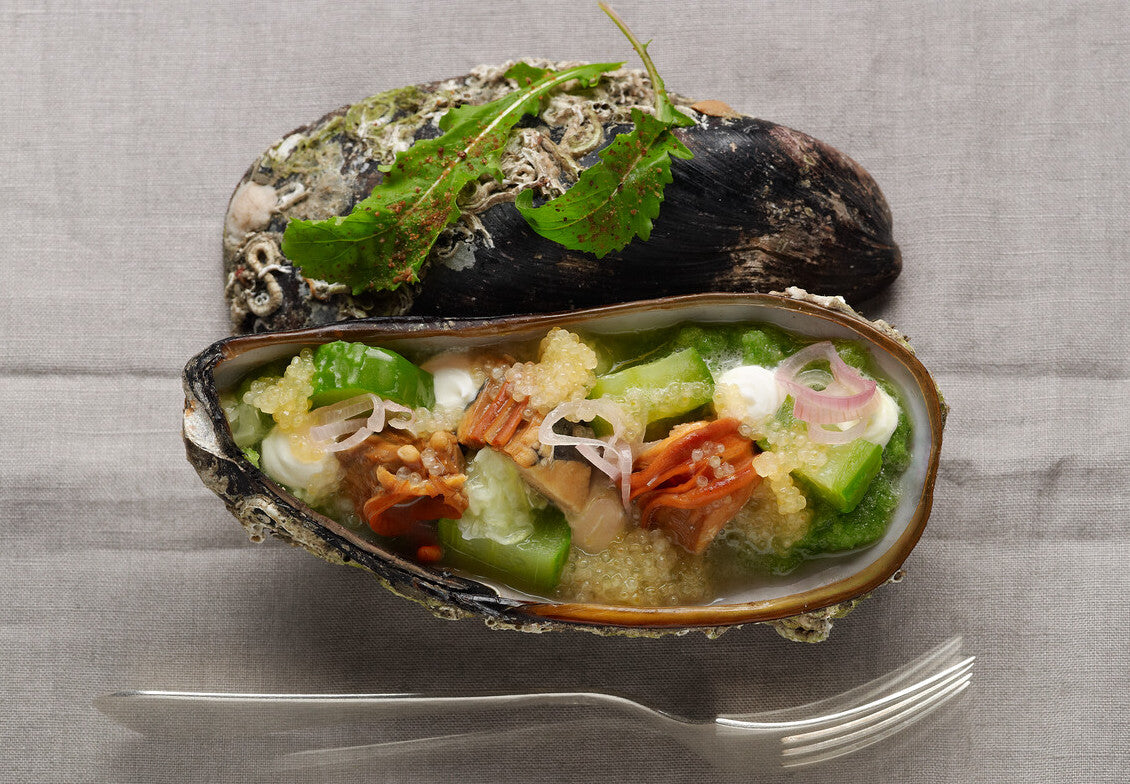 Eine vorbereitete Muschelschale mit einer Mischung aus Gemüse, Meeresfrüchten und Beilage, serviert auf einer grauen Tischdecke mit einer an die Seite gelegten Gabel.