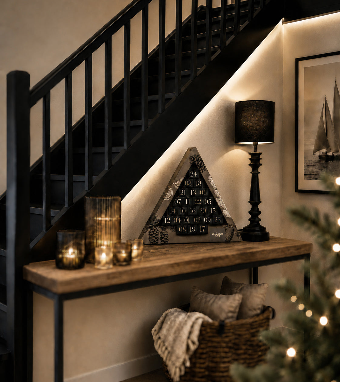 Auf einem Holztisch unter einer Treppe stehen Kerzen, der Sylter Adventskalender 2026 – ein dreieckiger Kalender der Sylter Manufaktur Johannes King, gefüllt mit hochwertiger Schokolade – und eine schwarze Lampe. Darunter steht ein Korb mit Decken. Ein gerahmtes Bild und ein Teil eines Weihnachtsbaums sind zu sehen, alles in warmes Licht getaucht.