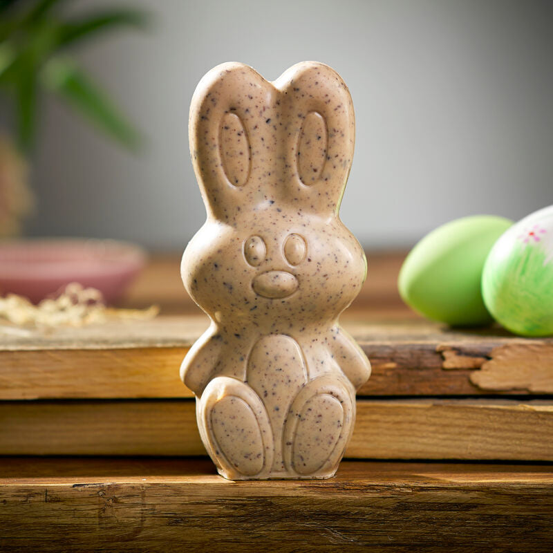 Eine gesprenkelte Seife in Hasenform namens Kings Schokoladen-Osterhase, Stracciatella von der Sylter Manufaktur Johannes King liegt auf einer Holzfläche. Im Hintergrund sind grün bemalte Eier und eine Pflanze mit grünen Blättern zu sehen. Diese Hasenseife erinnert an ein Ostergeschenk und hat geschnitzte Details für die Gesichtszüge und Ohren.