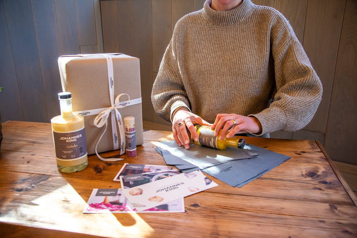 Eine Person in einem beigen Pullover sitzt an einem Holztisch und arrangiert Gegenstände, darunter Gewürzflaschen, Broschüren und eine verpackte Schachtel mit hochwertiger Geschenkverpackung der Sylter Manufaktur.