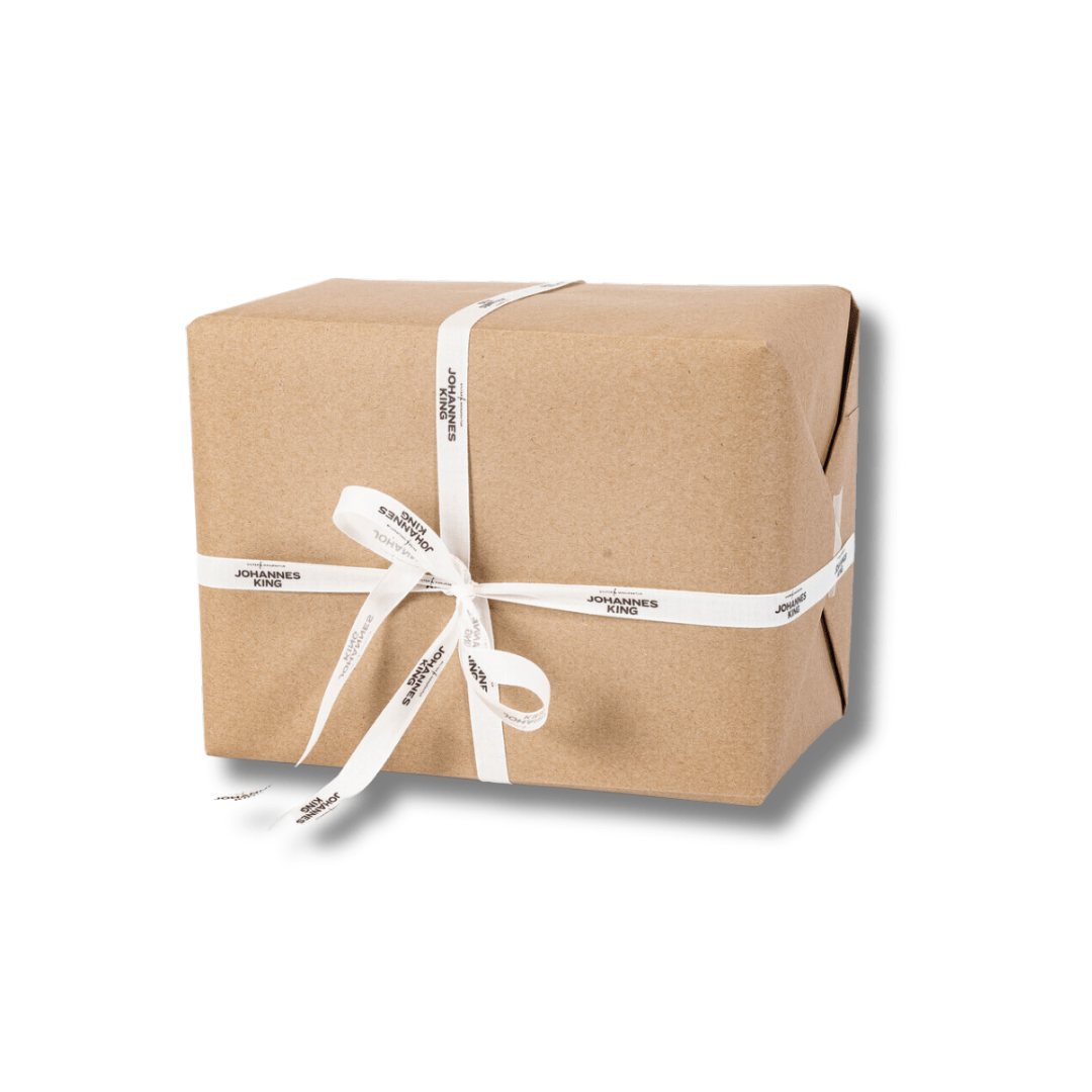 Ein rechteckiges Päckchen in braunem Papier ist mit einer weißen Schleife mit der Aufschrift „Sylter Manufaktur Johannes King“ verschnürt. Die hochwertige Geschenkverpackung ist schlicht und sauber verpackt und weist sichtbare Falten an den Ecken auf. Eine exklusive Notiz wertet das elegante Design auf einem schlichten weißen Hintergrund auf.