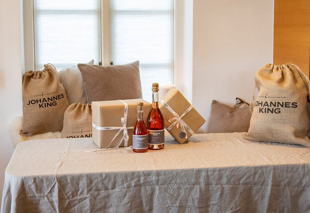 Geschenktüten und verpackte Geschenke der Sylter Manufaktur auf einem Tisch mit beiger Tischdecke, elegantem Design, einem Fenster im Hintergrund und zwei Flaschen Roséwein