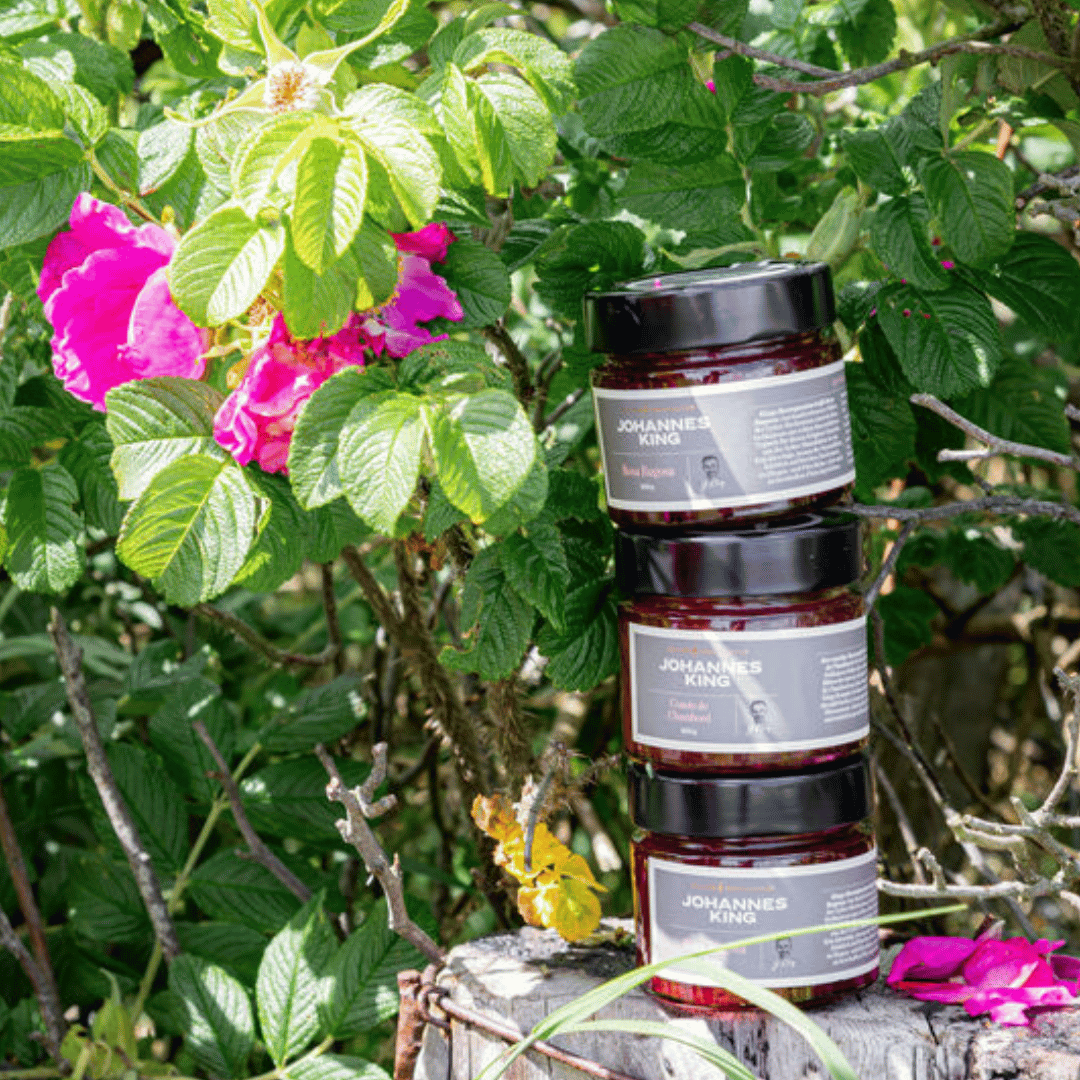 Drei Gläser Sylter Manufaktur Kings Rosa Rugosa Rosenaufstrich gestapelt auf einem Baumstumpf, umgeben von üppigem grünem Laub und leuchtend rosa Blüten.