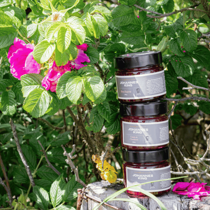 Drei Gläser Sylter Manufaktur Kings Rosa Rugosa Rosenaufstrich gestapelt auf einem Baumstumpf, umgeben von üppigem grünem Laub und leuchtend rosa Blüten.