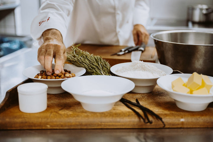 Ein Koch bereitet Zutaten auf einem Holztisch zu, mit Schüsseln mit Nüssen, Mehl, Butter und anderen Zutaten, die auf den Backprozess von Kings Haselnusskekse in einer professionellen Küche der Sylter Manufaktur hinweisen.