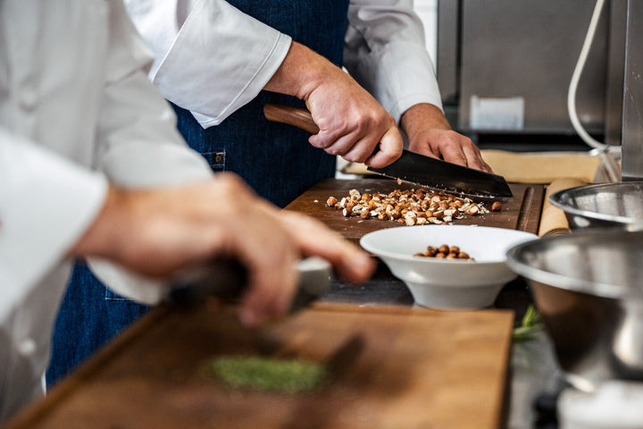 Ein Koch schneidet Haselnüsse auf einem Holzschneidebrett in einer professionellen Küche fein, während in der Nähe andere Zutaten für Kings Haselnusskekse zubereitet werden.