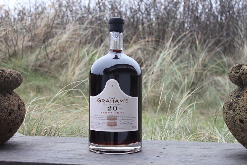 Eine Flasche Graham's Tawny Port 20 Jahre 4,5l steht auf einer Holzoberfläche mit Gras und Laub im Hintergrund.