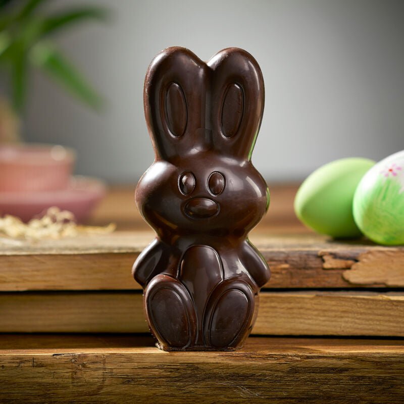 Ein Kings Schokoladen-Osterhase, dunkle Schokolade wird auf einer Holzoberfläche präsentiert, umgeben von Ostereiern und einer Pflanze im Hintergrund.