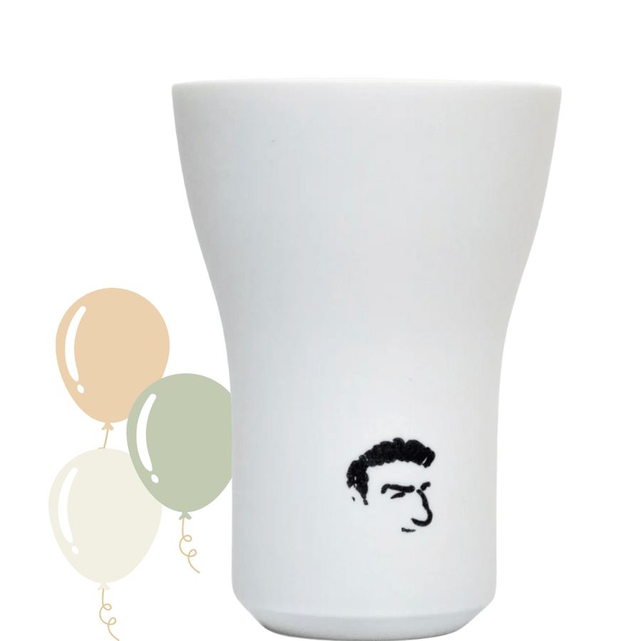 Eine weiße Keramikvase mit einer einfachen schwarzen Strichzeichnung eines Gesichts darauf, begleitet von einem Trio aus Luftballons in gedämpften Farben auf der linken Seite. Bei dem Produkt handelt es sich um den Kings Hering Berlin Becher *King Sonderedition* der Sylter Manufaktur.