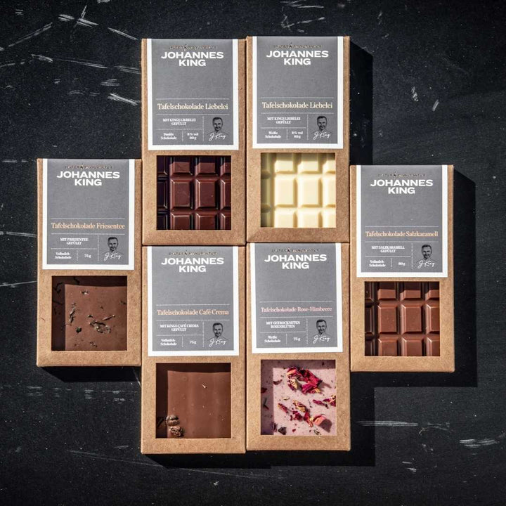 Neun sortierte Sylter Manufaktur Kings Liebelei Tafelschokoladen dunkel, jeweils mit unterschiedlichen Geschmacksrichtungen und Kakaoanteilen, übersichtlich in drei Reihen auf einer dunklen, strukturierten Oberfläche angeordnet.