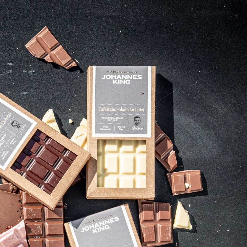 Eine Auswahl an Kings Liebelei Tafelschokoladen-Hellriegeln und -Stücken, darunter weiße Schokolade, auf einer dunklen Oberfläche mit der Marke „Sylter Manufaktur“.