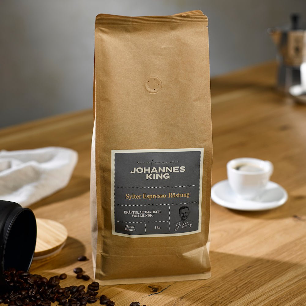Eine Packung Sylter Manufaktur Kings Espresso groß steht auf einer Holzoberfläche, im Hintergrund sind eine Tasse Espresso, ein Deckel und lose Kaffeebohnen zu sehen.