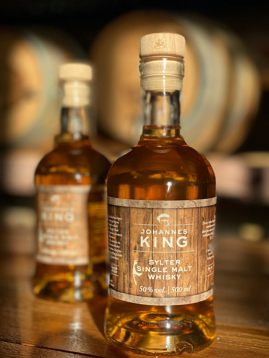 Zwei Flaschen Sylter Manufaktur Kings Sylter Single Malt Whisky, 50 % Vol., 500 ml, prominent präsentiert vor Holzfässern.