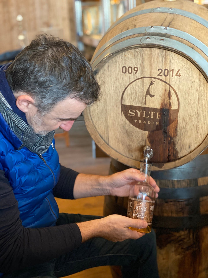 Ein Mann in einer blauen Weste begutachtet eine Flasche Kings Sylter Single Malt Whisky neben einem Holzfass mit der Jahreszahl 2014.