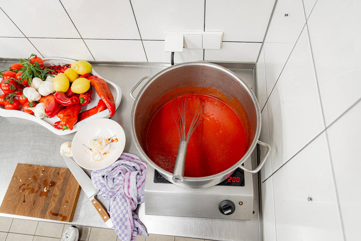 Ein großer Topf mit Tomaten und Kings Sylter Feuer – scharfe Chili-Sauce mit einem Schneebesen darin steht auf einer Kochfläche, daneben ein Tablett mit frischen Zutaten wie Tomaten, Zitronen und Kräutern.