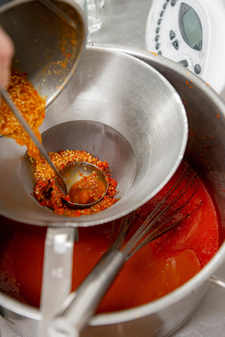 Gemixte Chilimischung durch ein Sieb in eine Metallschüssel gießen, im Hintergrund Schneebesen und Küchengeräte, um Kings Sylter Feuer zuzubereiten – scharfe Chili-Sauce als Tabasco-Alternative von der Sylter Manufaktur.