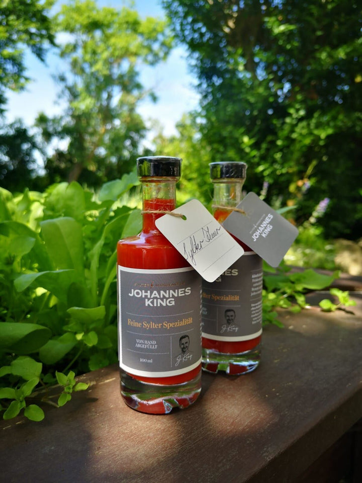 Zwei Flaschen Sylter Manufaktur Kings Sylter Feuer – scharfe Chili-Sauce mit Produktetiketten, platziert auf einer Holzoberfläche im Freien mit grünem Blattwerk im Hintergrund.