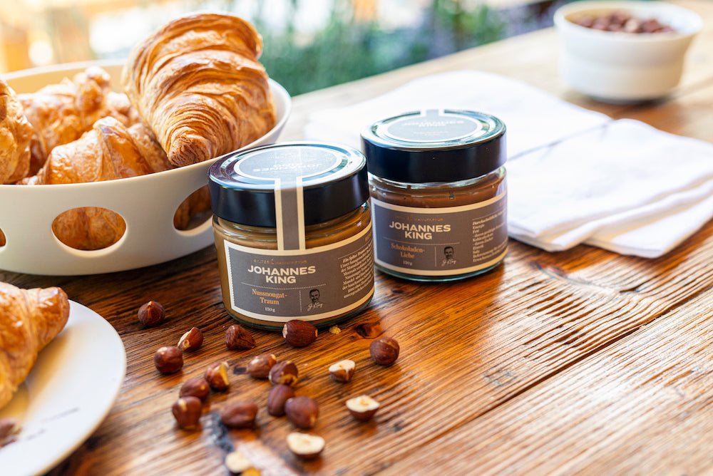 Eine Auswahl frisch gebackener Croissants serviert auf einem Holztisch mit zwei Gläsern Aufstrichen der Marke „Kings Nussnugat – Traum mit Sylter Meersalz“ der Sylter Manufaktur und Haselnüssen.