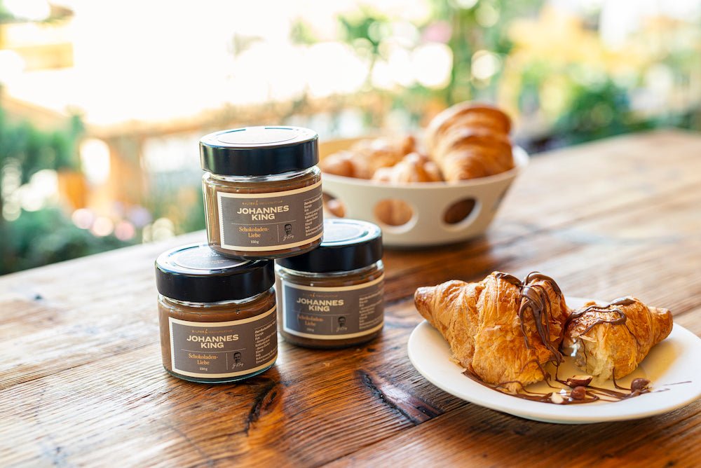 Drei Gläser Sylter Manufaktur-Marmelade sowie zwei Croissants, eines davon mit Kings Nussnugat – Traum mit Sylter Meersalz beträufelt, auf einem Holztisch.