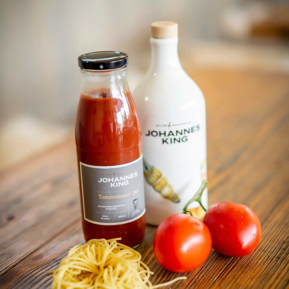 Auf einem Holztisch stehen zwei Flaschen Kings Pure Sylter Tomatensauce neben einem Paar frischer Tomaten und einem kleinen Nest ungekochter Nudeln.