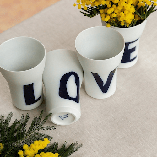 Vier Hering Berlin Letter Cups mit Buchstaben darauf, die das Wort „Love“ ergeben, auf einem Tisch mit gelben Blumen und Grün im Hintergrund.