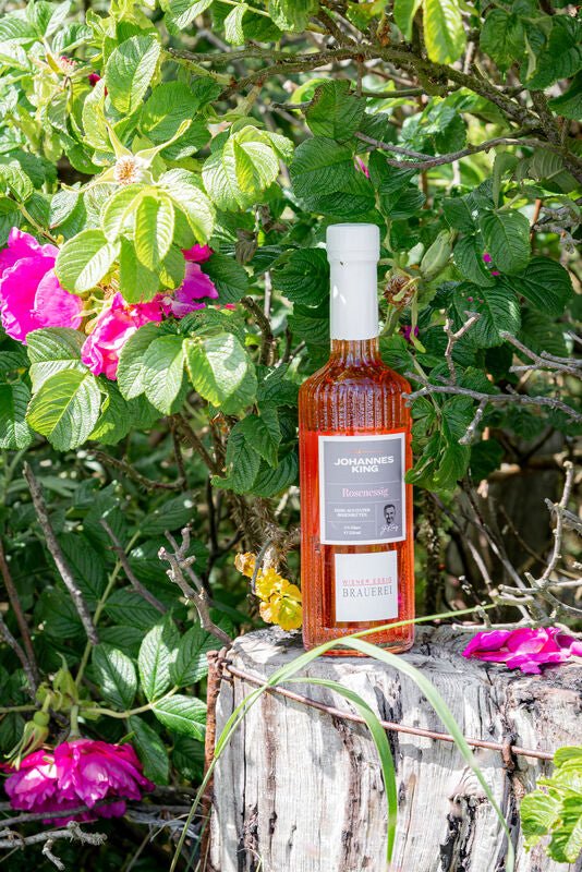 Eine Flasche Kings Sylter Rosenessig von der Sylter Manufaktur steht auf einem rustikalen Baumstumpf, umgeben von grünem Laub und rosa Blüten.