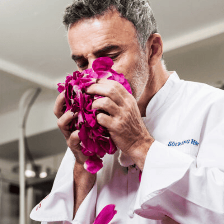 Ein Mann in einem weißen Kochmantel hält einen großen Strauß rosafarbener Blütenblätter in der Hand und riecht intensiv daran, was an den einzigartigen Duft des Königs Sylter Rosengin erinnert.